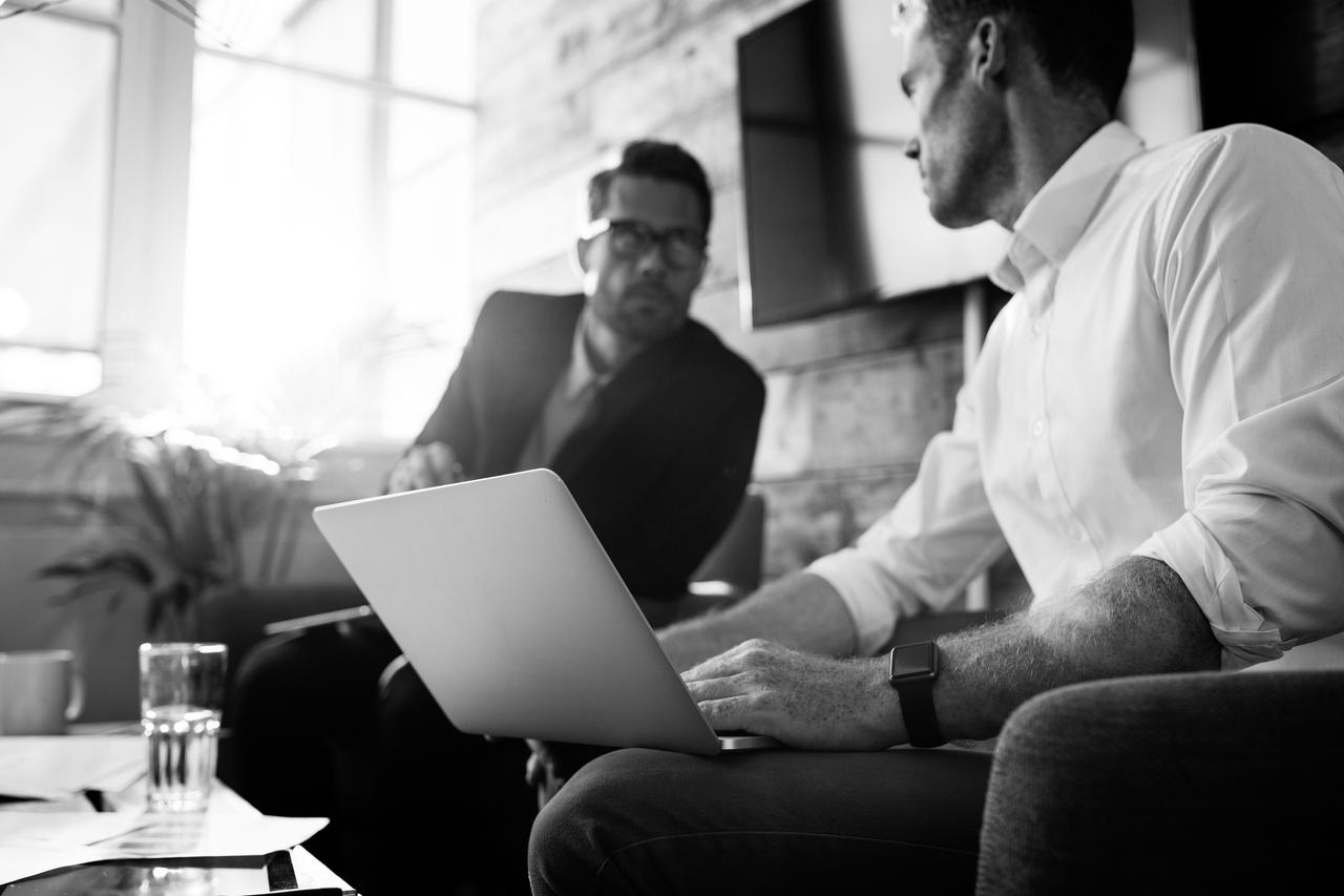 two men having a conversation on a couch with one holding a laptop