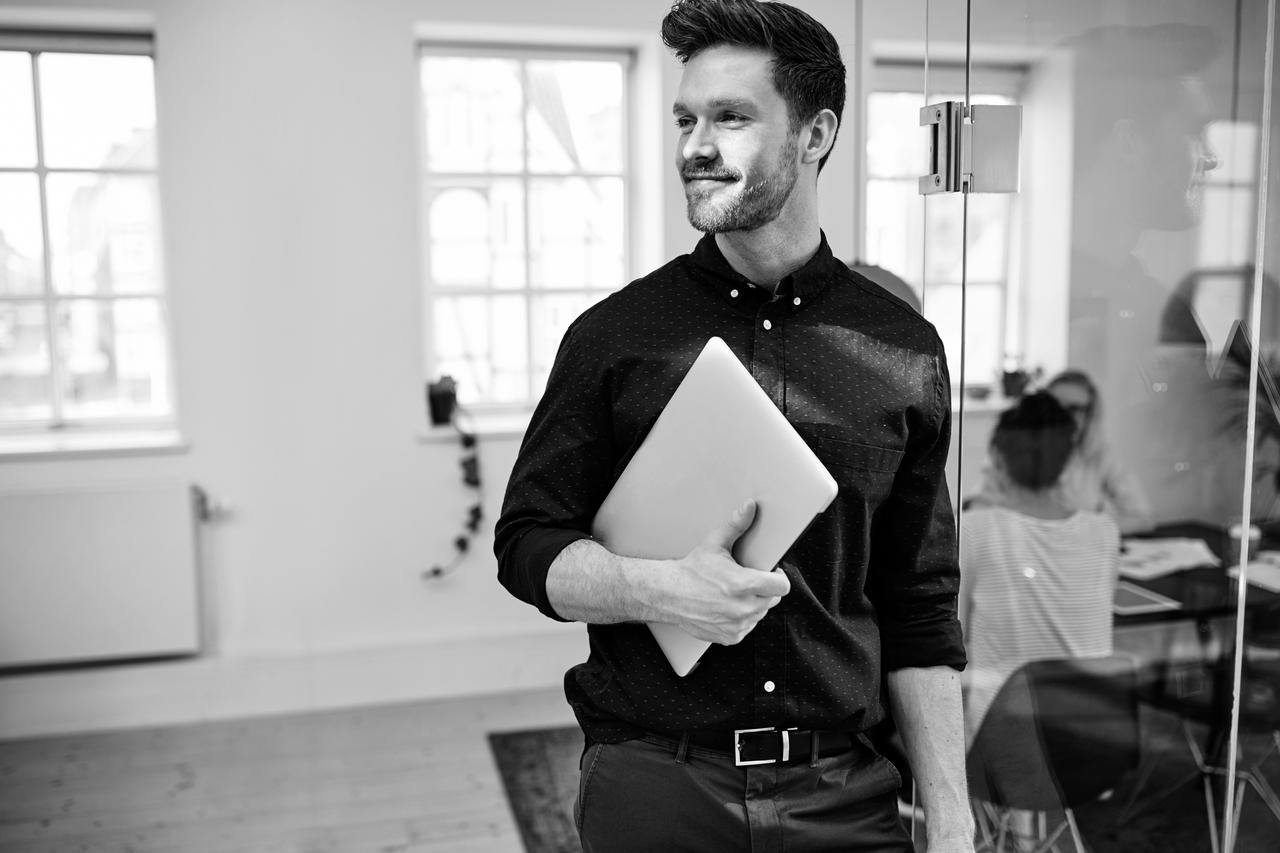 Smiling young businessman walking with his laptop in a modern office with colleagues at work in the background