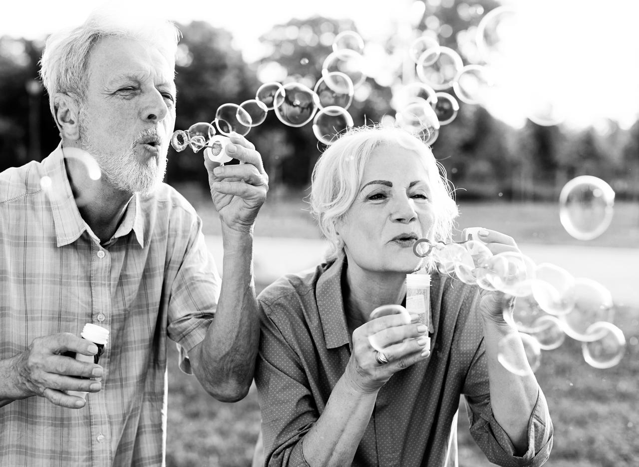 Happy active senior couple having fun blowing soap bubbles in park outdoors.