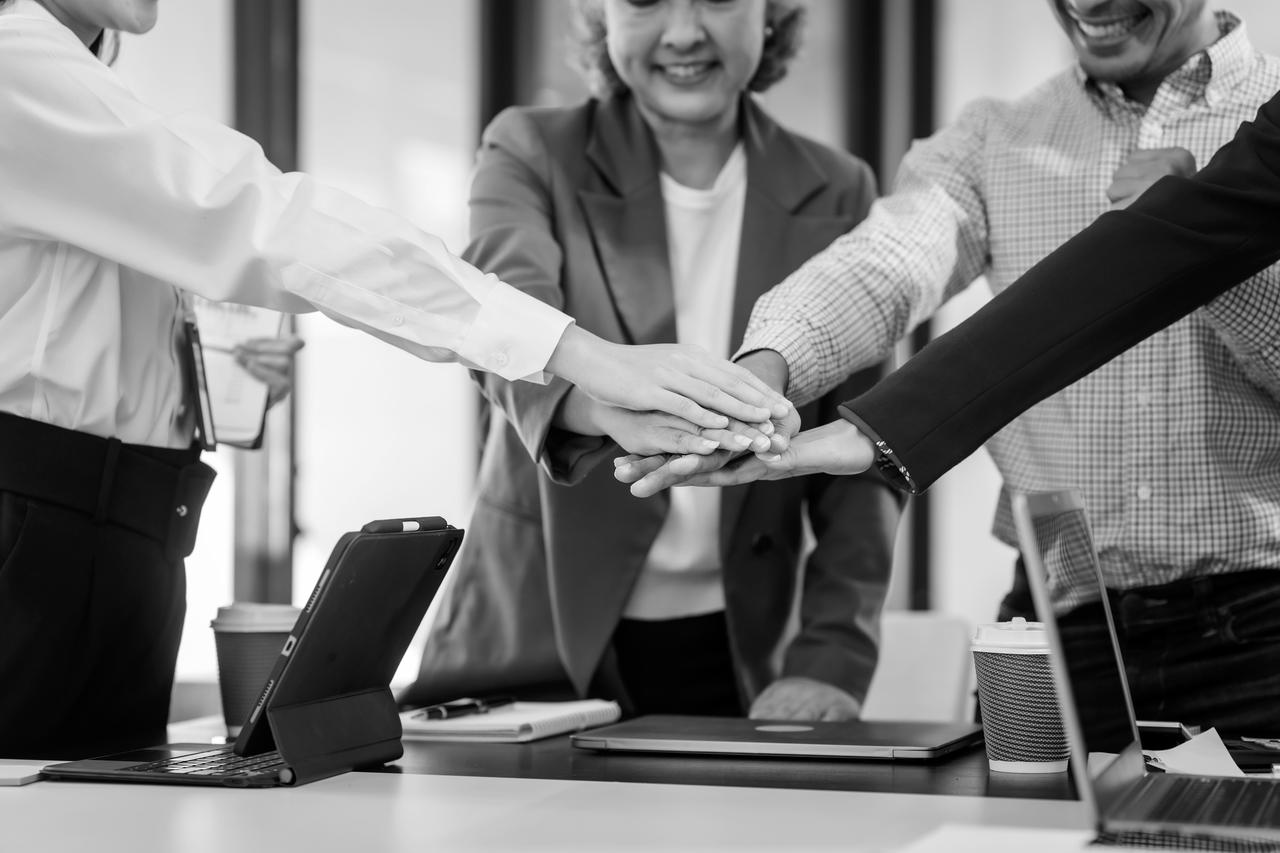 Four people in an office put their hands together as a team. They smile and wear work clothes. laptops, coffee, and papers on the table.