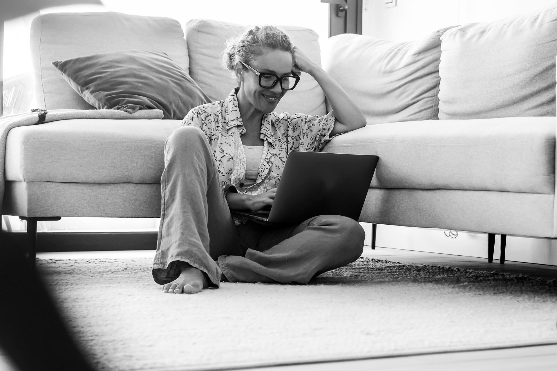 Carefree modern adult woman using laptop at home sitting on the floor against a grey sofa