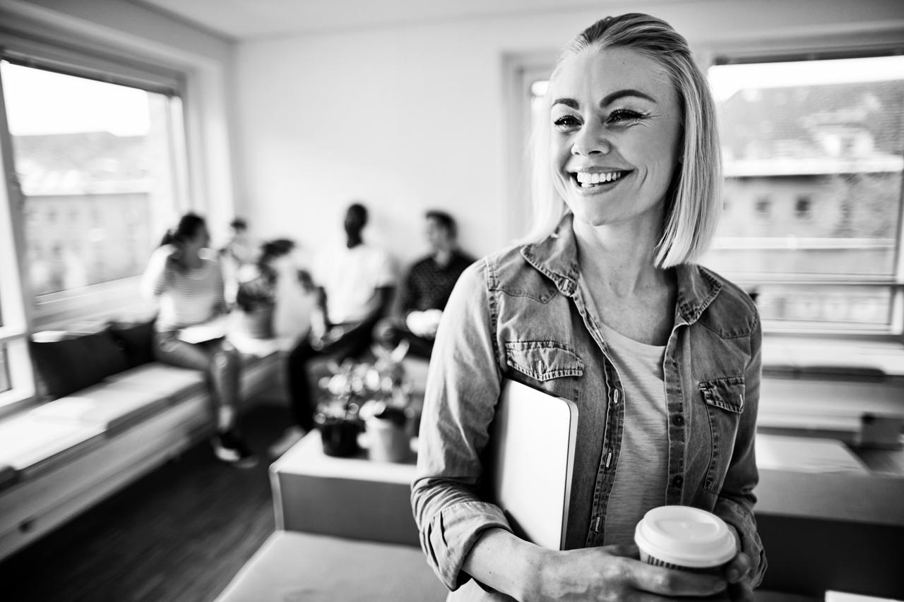 Young woman smiling, carrying a laptop and holding a coffee in an open plan office or shared workspace